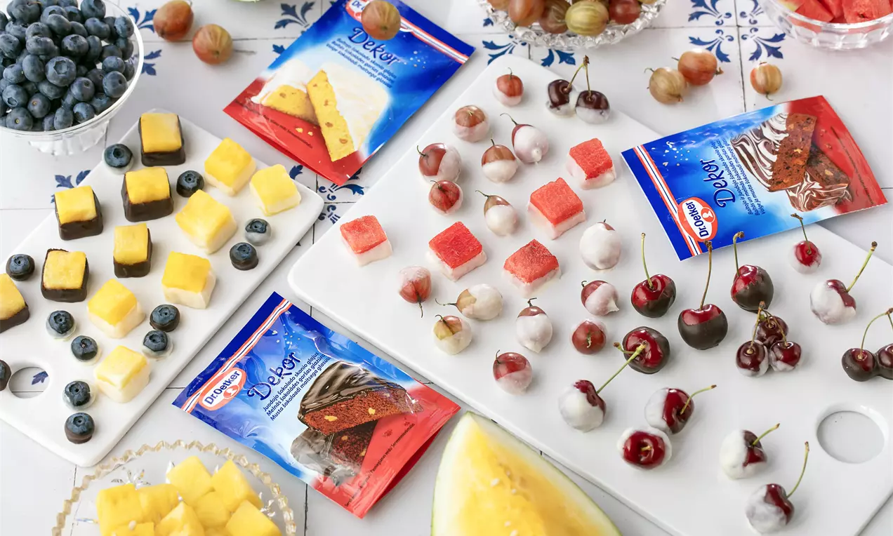 Uz balta galda ar zilu ziedu rakstu izkārtoti krāsaini augļi un ogas, tostarp ķirši, mellenes, ērkšķogas, arbūzs un ananasi, blakus saldumu iepakojumiem.