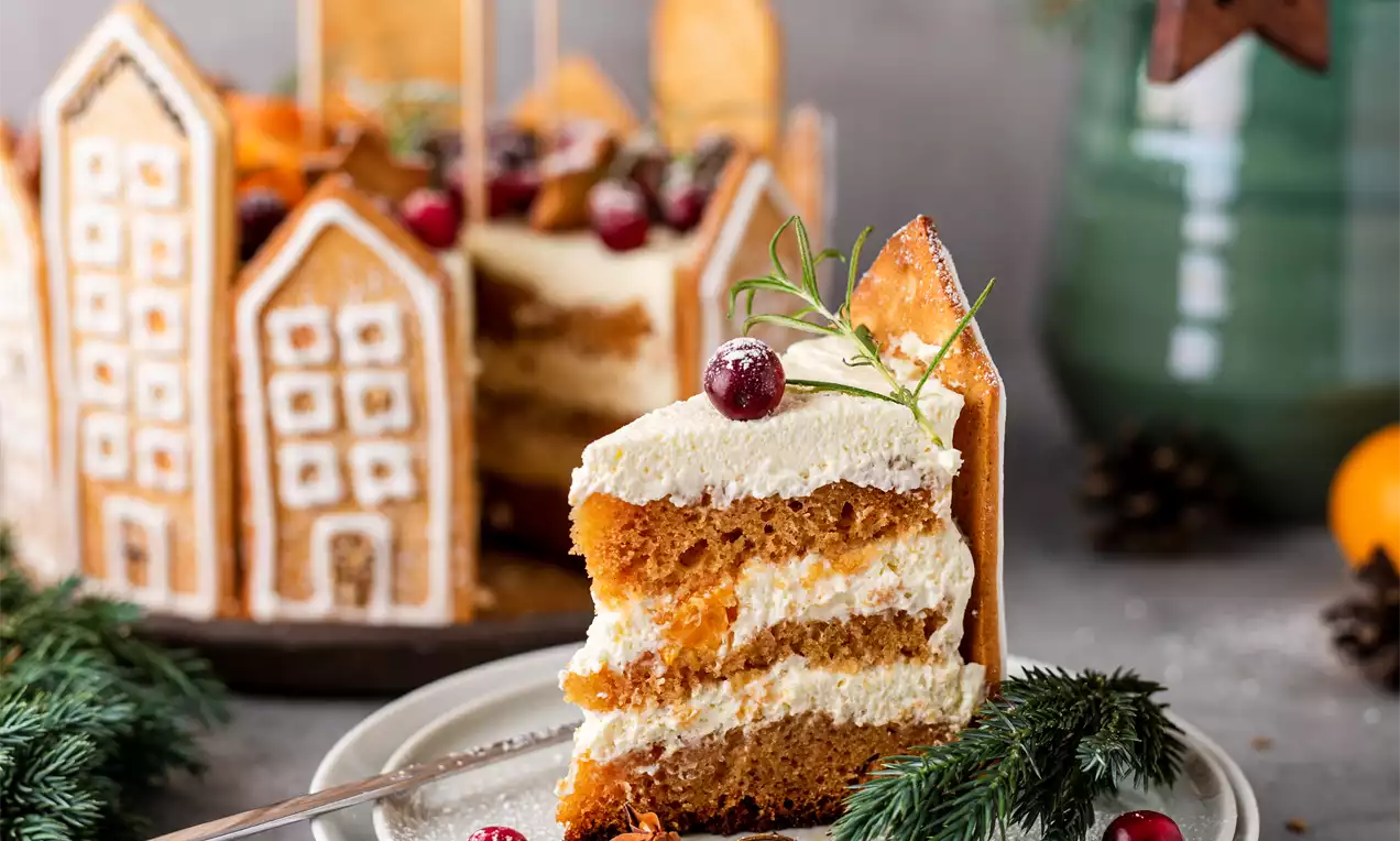 Apelsīnu tortes trīsslāņu griezumā redzami kupli biskvīta un krēma kārtu izslāņojumi, dekorēts ar dzērvenēm, kanēļa standziņu un egles zariņu